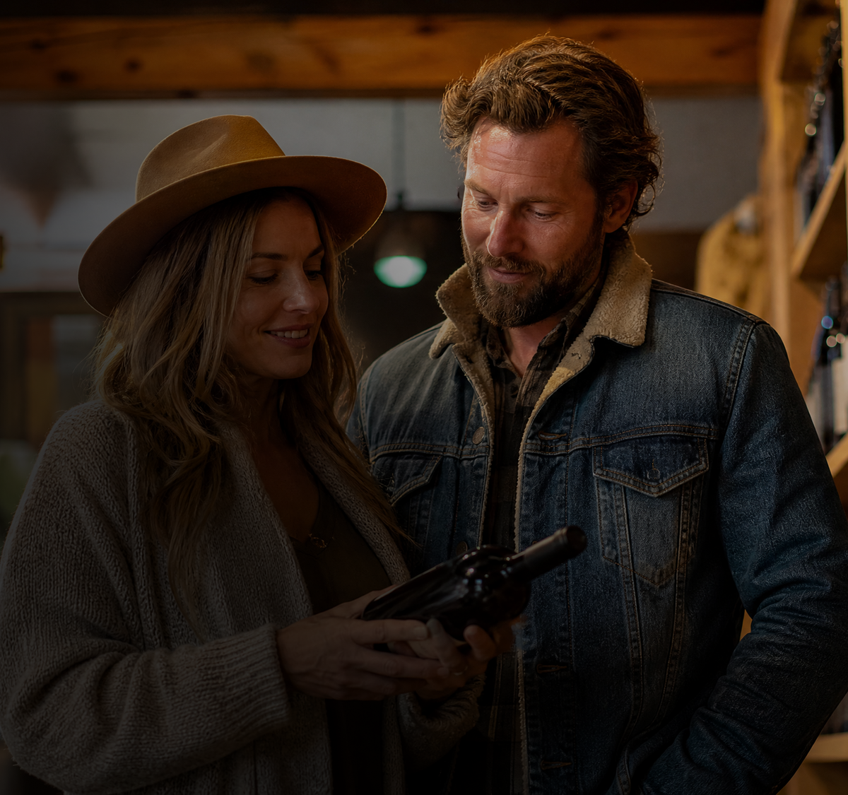 A man and a woman standing together in a rustic indoor setting, looking at a dark bottle held by the woman. The woman wears a tan wide-brim hat, a taupe knit cardigan, and a dark top.