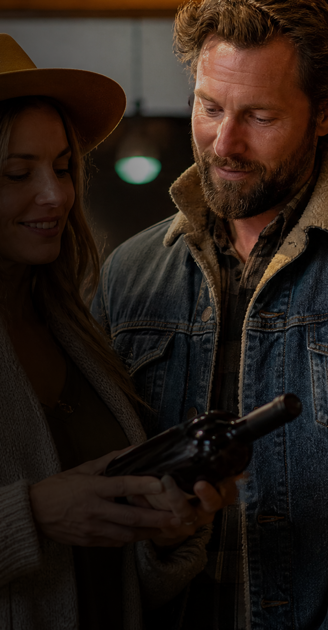 A man and a woman in a dimly lit indoor setting look closely at a dark wine bottle held by the woman. The man wears a denim jacket with a shearling lining, and the woman wears a wide-brimmed hat.